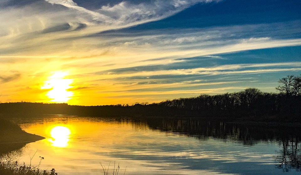 Chickasaw National Recreation Area The sun setting low on the horizon, shining through long, wispy clouds and reflected on a lake lined by trees and shrubs.