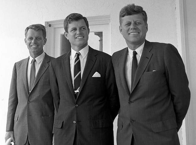 A black and white photo.  From left to right, Bobby, Ted, and John Kennedy.  All wearing dark suits standing in front of a white wall.