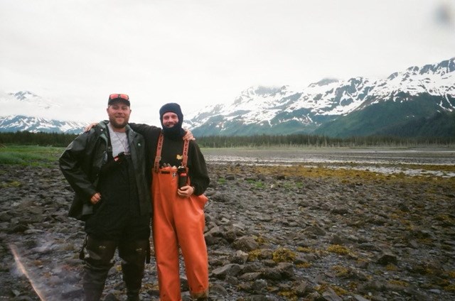 two white mails in outdoor gear posing in front of a field with mountains in the background