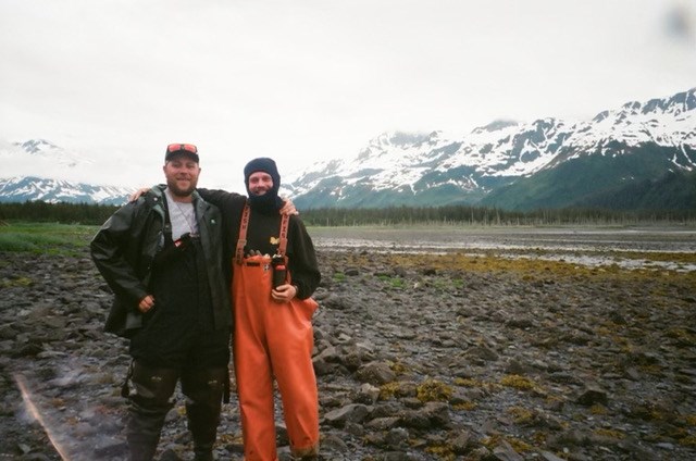 Intern Matin Schneider and seasonal Archeology Technician Sean Sullivan at Surprise Bay visiting Kenny Fox Mine. two white mails in outdoor gear posing in front of a field with mountains in the background