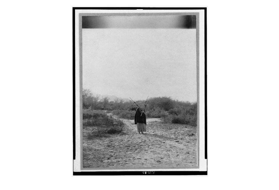 Historic photograph of an O'odham woman with a kiaha on her back, walking away.