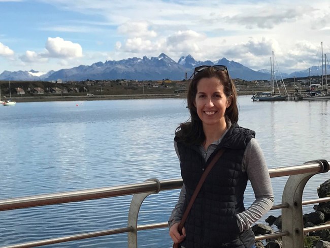 Woman smiling, standing next to metal railing with a large body of water in the background.