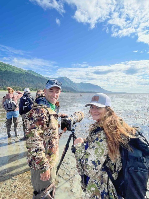 thumbnail_Brandon and Summer profile A man and woman wearing camouflage jackets and grey ball caps stand in front of a camera on a tripod standing on mudflats. Mountains covered with trees in the distance.