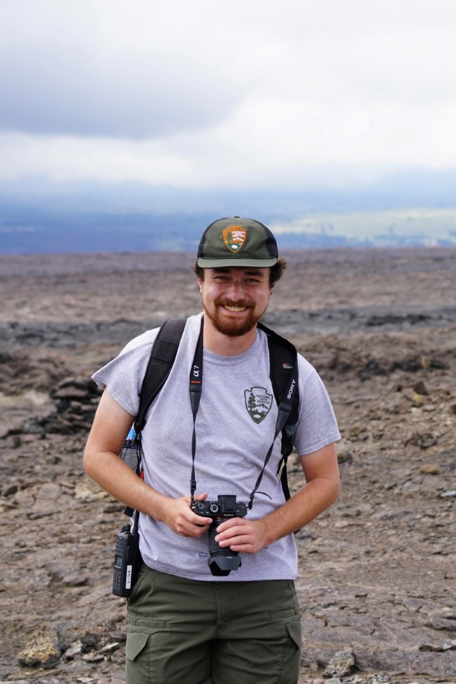 Daniel in action Man with NPS uniform, backpack, gps unit, and binoculars standing in front of a Hawaii landscape