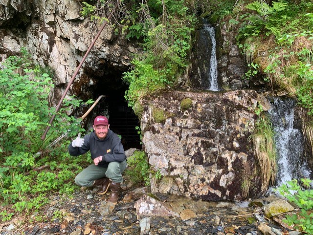 Intern Martin Schneider in-front of upper adit at at Glass-Heifner Mine. White man kneels down and throws a thumbs up next to a mine entrance surrounded by rocks, plants, and a small stream