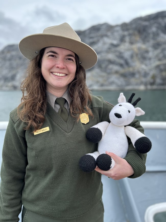 A park ranger smiling and holding a crocheted mountain goat in her arms.