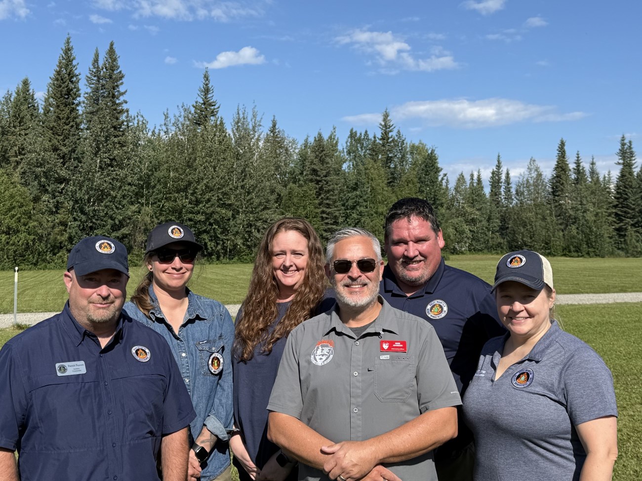 A group of six people wearing similar shirts and hats with an Incident Management Team logo smile at the camera; trees in the background.