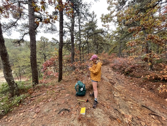 Young female scientist stands in fall forest with backpack and measuring tape at her feet.