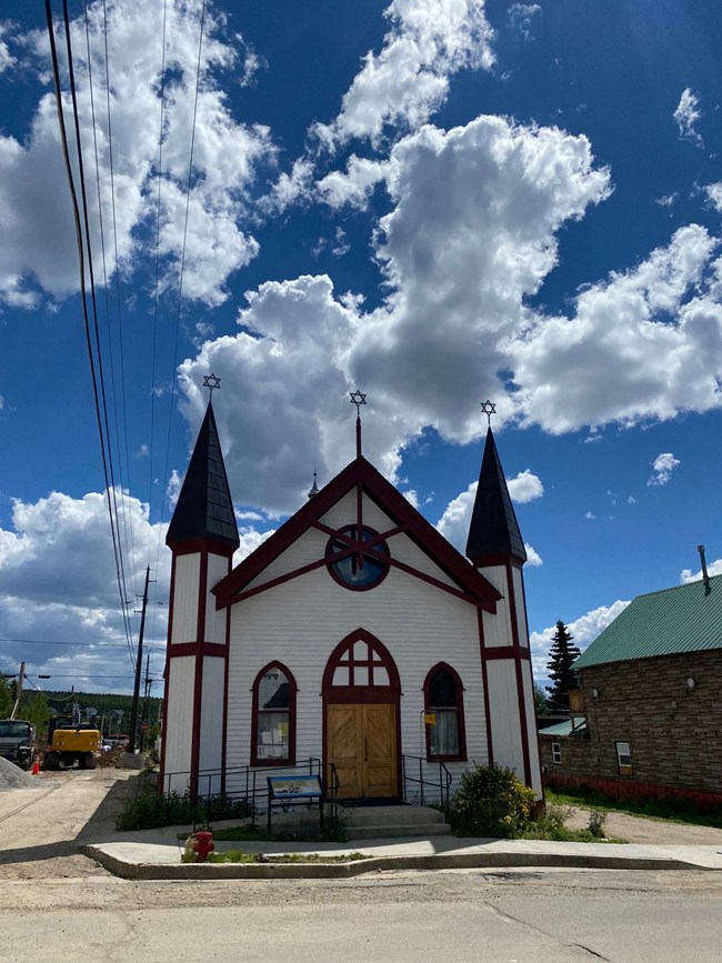 View of the main façade of the historic frame synagogue building with steeply pitched roof flanked by two small towers under a dramatic blue sky with clouds.