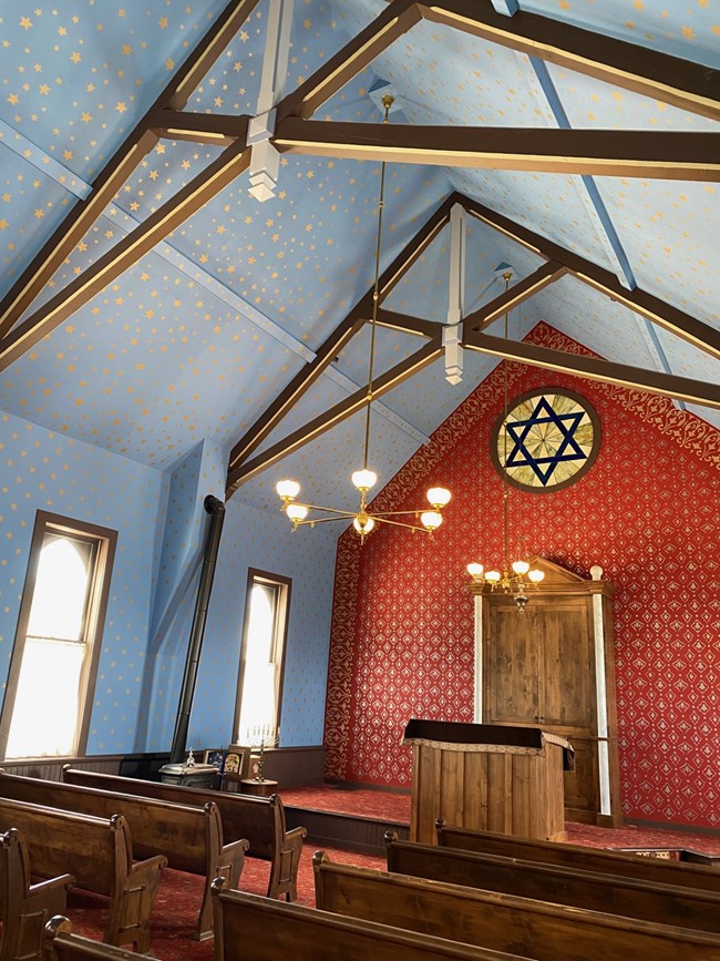 Interior view of the sanctuary (museum) space showing a painted blue ceiling with gold stars and open trusswork. Red wall treatment and a circular stained-glass window depicting the Star of David serve as the backdrop behind the bimah.