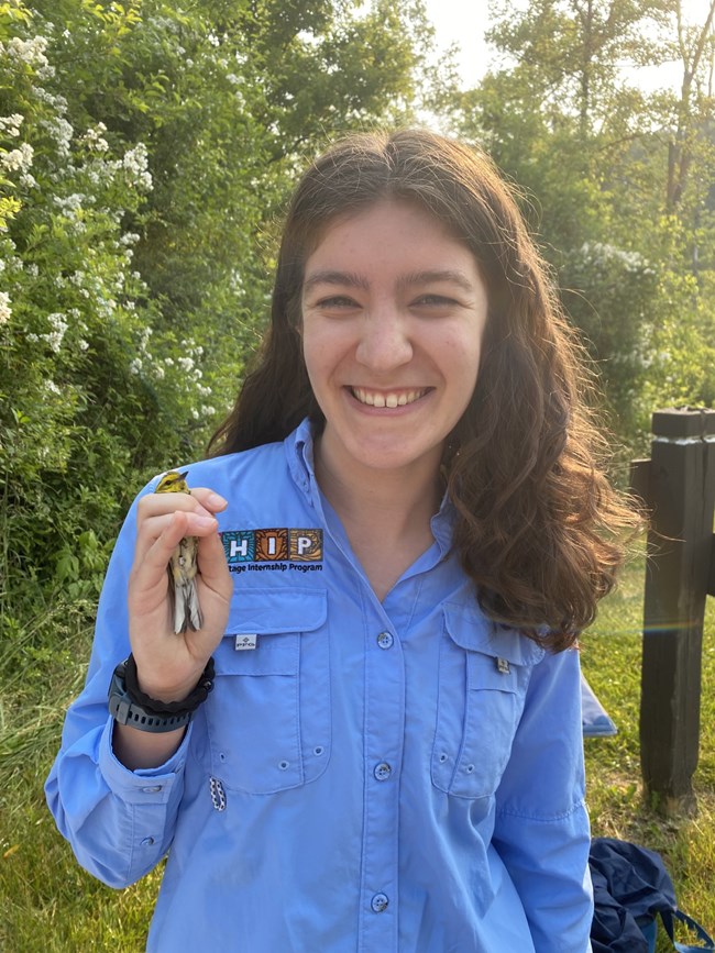 A woman in a blue shirt with Latino Heritage Internship Program logo smiles and holds a small yellow bird.