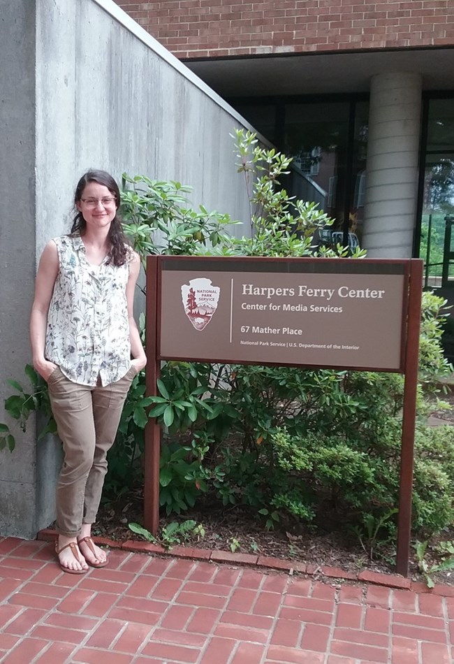 Perri Meldon stands outside the Harpers Ferry Center (HFC) in West Virginia. White woman stands outside the Harpers Ferry Center (HFC) in West Virginia. She wears a sleeveless floral blouse, khaki pants, and sandals.