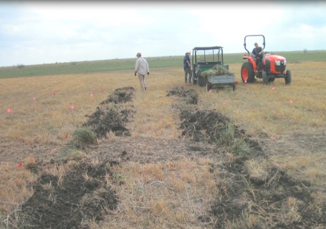 Personnel with a tractor and other equipment on a field with red flags and trenches.