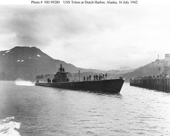 Dark submarine above water with people standing on deck and on the dock, with mountains in background.