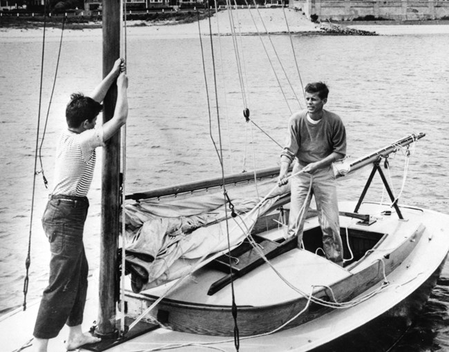 A black and white photo.  Ted Kennedy is pulling a rope while Jack Kennedy watches.  They are inside a sail boat on the water