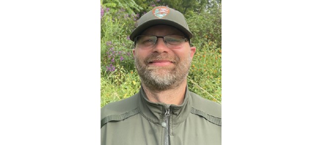 Caleb Tzilkowski Tzilkowski smiling outside in front of lush greenery in an NPS field uniform and glasses.
