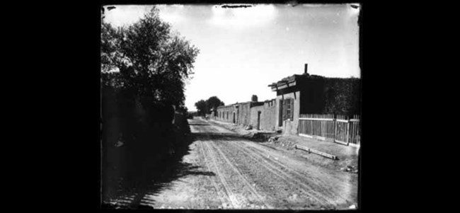 lt= A historic picture of a dirt road lined with adobe buildings.