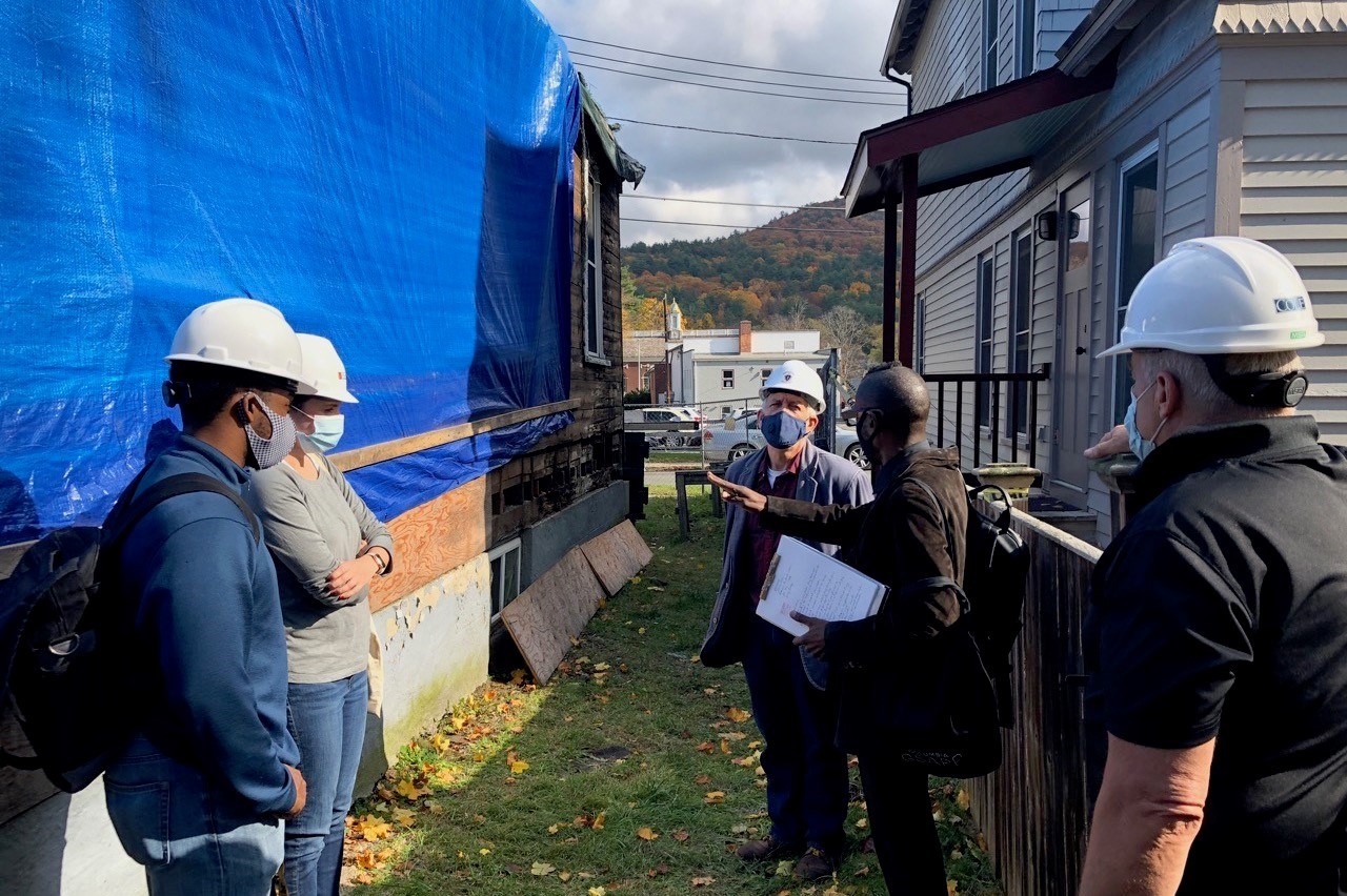 Clinton Church Restoration Five people stand in discussion between two building; building at left covered in construction tarp. Four people wear hardhats, all wear surgical masks.