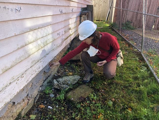Clinton Church Restoration Person wearing hardhat crouches on grass next to base of building, outstretching hand to feel very worn cement or stone foundation below weathered white siding.