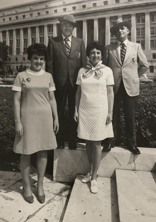 Two men in blazers, a woman in a beige dress, and woman in polkadot dress