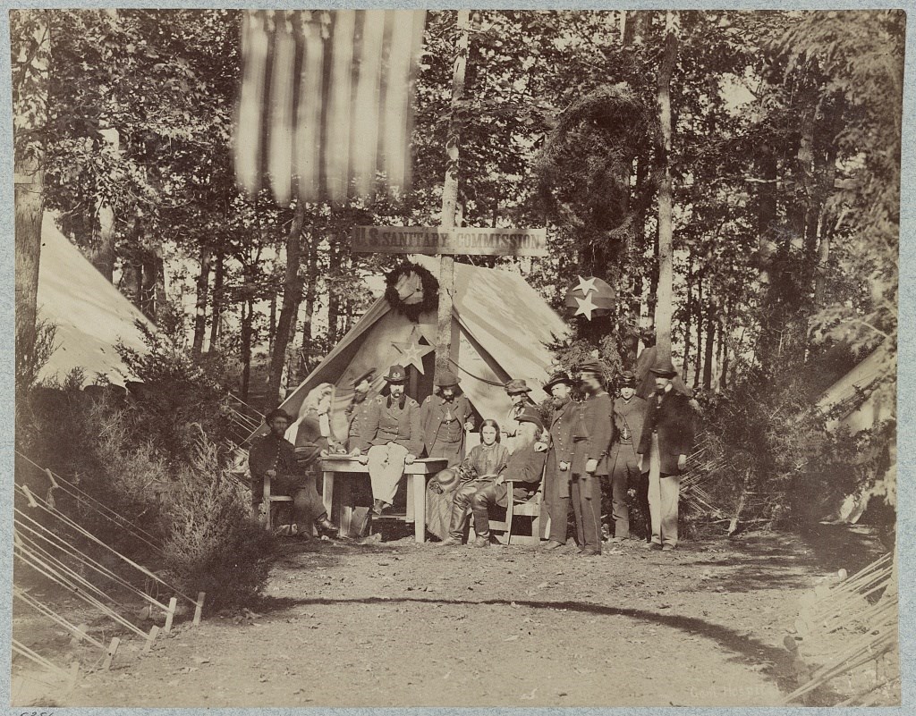 Black and white photograph of group of white tents in forest with the sign U.S. Sanitary Commission. There are men standing, seated in a chair, and seated atop a table, but all wears the dark army uniform. There are also two women in dark dresses.