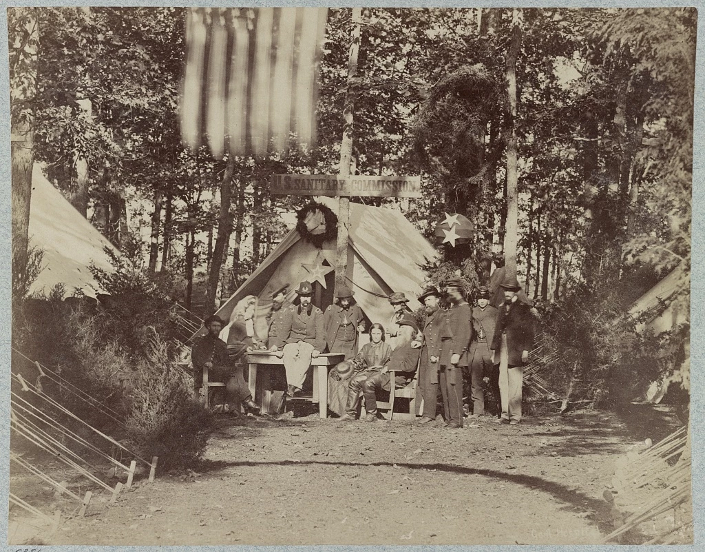 ussccamp Black and white photograph of group of white tents in forest with the sign U.S. Sanitary Commission. There are men standing, seated in a chair, and seated atop a table, but all wears the dark army uniform. There are also two women in dark dresses.