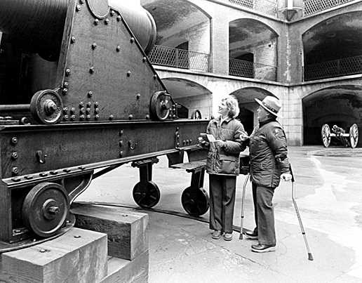 Vicky White, in uniform and walking with arm crutches, talks to a visitor standing next to a large cannon.