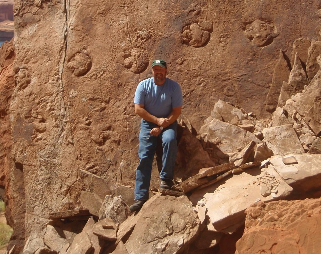 Vincent Santucci a man with a beard and ball cap stands next to a large boulder with a track of dinosaur footprints clearly visible