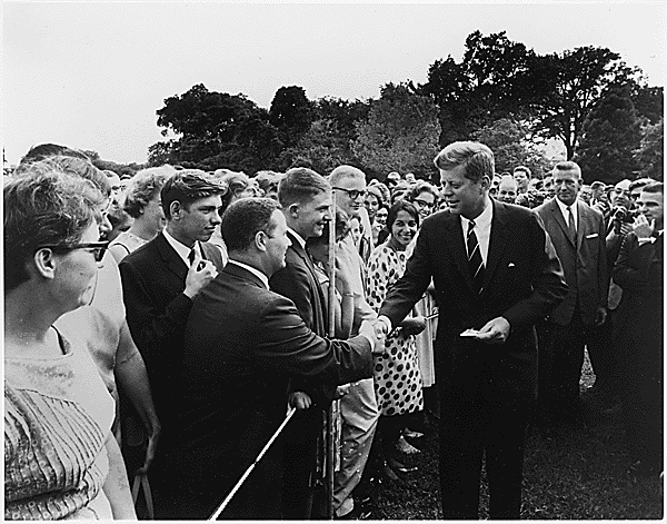 A crowd of youth on the left shake hands with President Kennedy on the right.  The photo is black and white.