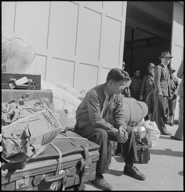 A man sits among stacks of luggage outside while other men, including soldiers, look toward the viewer from the background