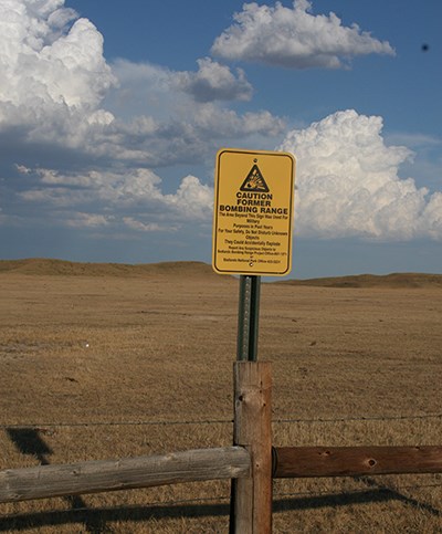 a yellow sign on a wooden fence in front of rolling brown prairie reads "caution former bombing range"