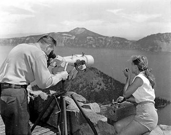 A man looks through a telescope out over the water.  A woman wearing shorts talks into a hand held radio.