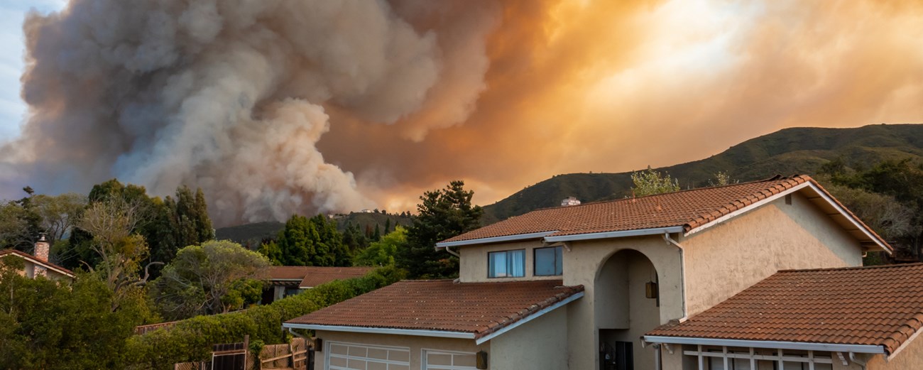 wildfire smoke billows from a fire on a hill above a group of houses