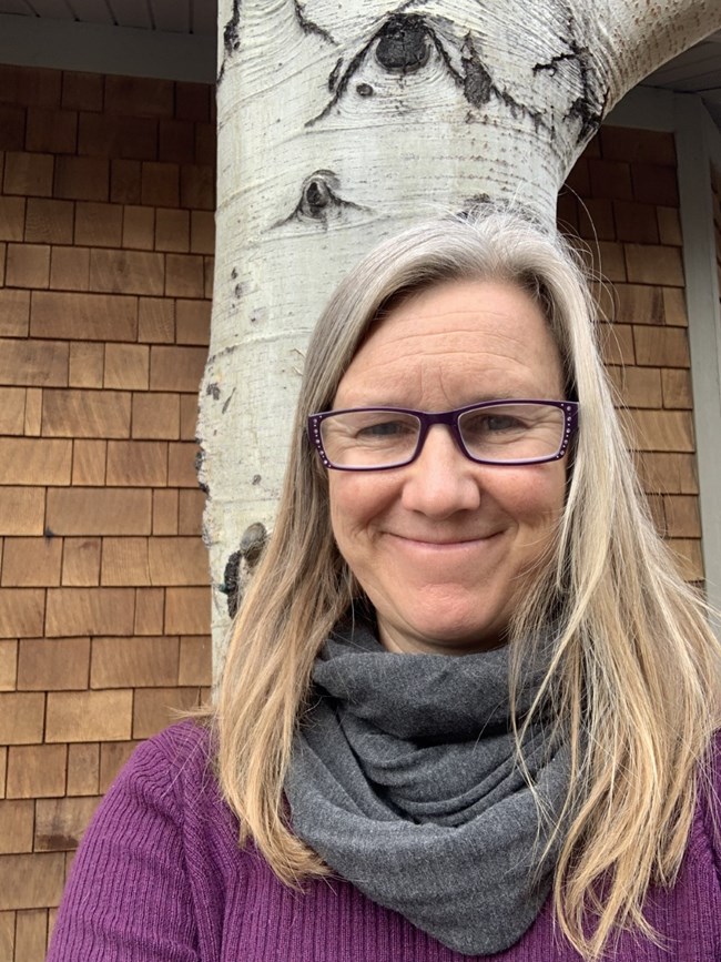 woman stands in front of a tree in front of a brick building and looks directly at the camera