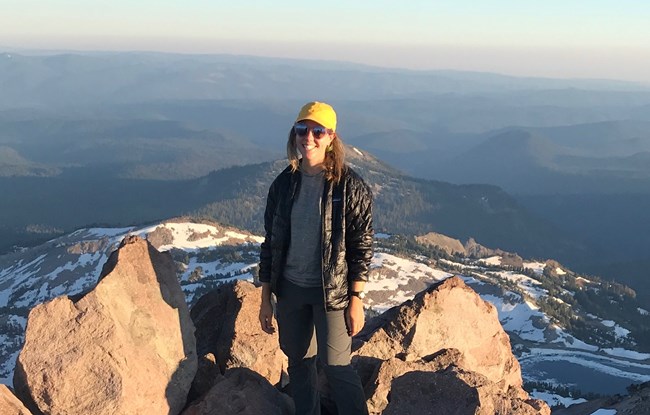 woman stands alone in a rocky outcrop facing the camera with a huge expanse of forested land behind her