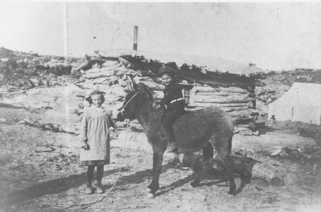 black and white image of a young boy in a sailor outfit sitting astride a shaggy burro. a girl in a pinafore holds the bridle.  in the background is a rough log cabin, a canvas tent, and a rocky backdrop
