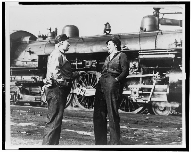 Two women in work clothes laugh together in front of a locomotive
