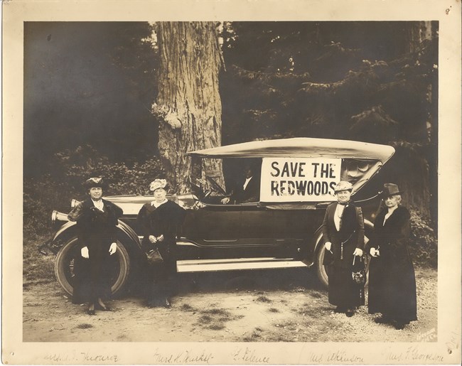 Women stand in front of an old fashioned with with a sign that reads 'Save the Redwoods'. These women were a part of the movement that helped preserve redwoods across the state of California.