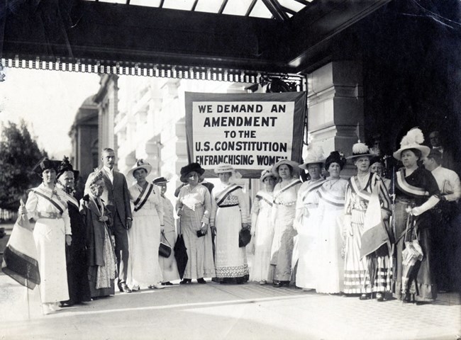 A historical photo of a group of women in long dresses, standing in front of a sign that demands women's suffrage.
