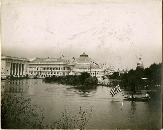 Black and white of body of water with small boat across it with USA flag and large white buildings in the background.