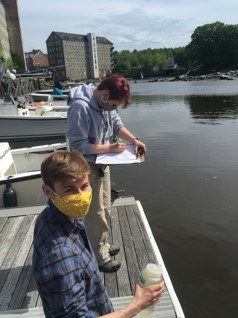 Two students in masks stand on a dock taking water quality samples