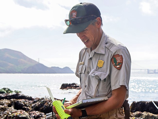 Ecologist in NPS uniform smiles down at a clipboard against the backdrop of rocky intertidal habitat.