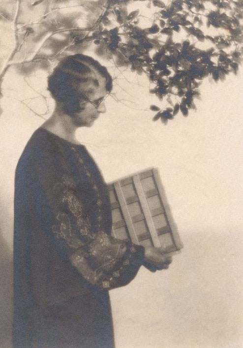 sepia toned photo of a woman holding a square wood object of some kind, standing under a tree