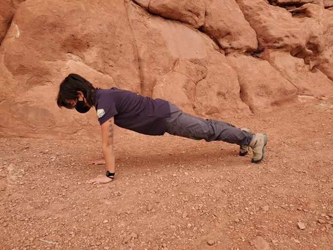 A female wearing a navy shirt and grey pants is poised in the plan position. Behind her is an orange colored rock wall