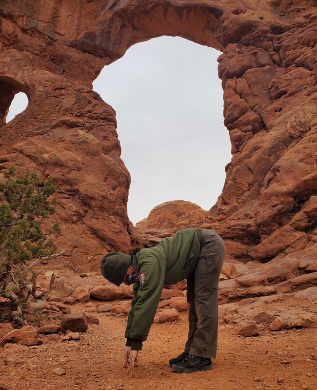 A female park ranger in uniform stands bent over at the waist with her fingertips touching the ground. Behind her is a large orange colored natural rock arch.