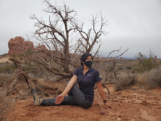 A female wearing a navy shirt and grey pants sits on the ground twisting her torso. Behind her is a juniper tree and a landscape of tan rocks and green shrubs.