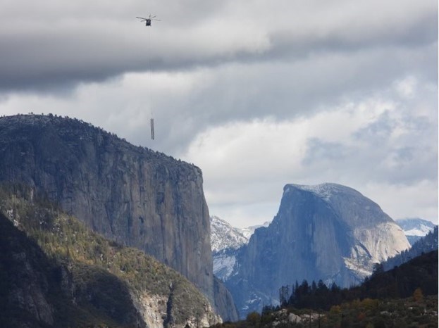 A helicopter hoisting a powerline tower flies in front of a mountain range