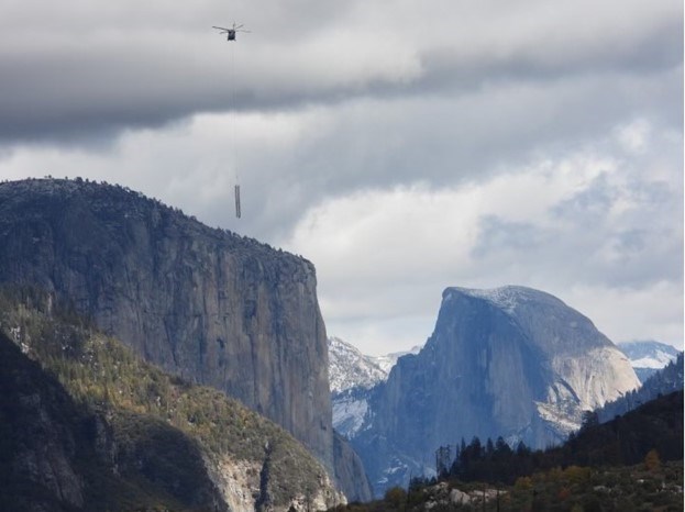 lt= A helicopter hoisting a powerline tower flies in front of a mountain range