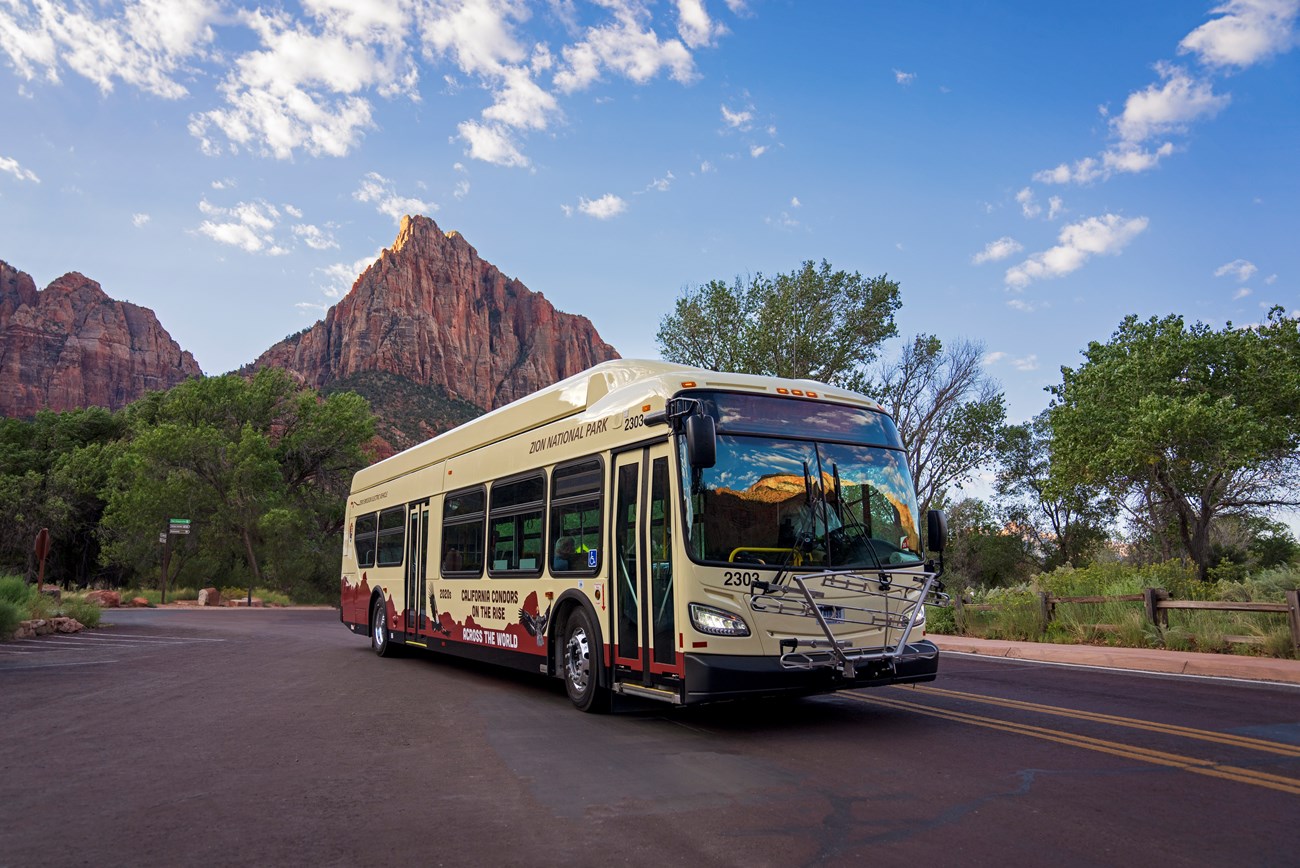 A beige shuttle bus on a dark asphalt road in front of a large, triangular sandstone mountain.