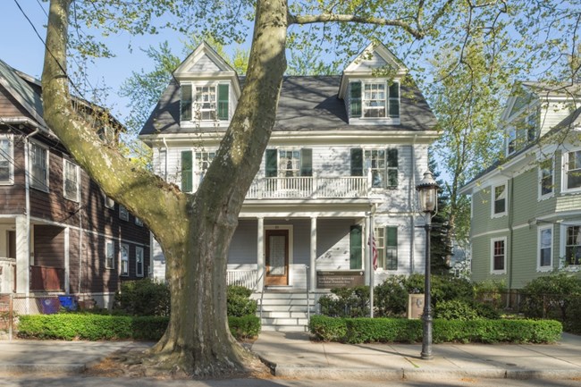 Grey house on a tree-lined street. US Flag on pole. Large tree in front of house.